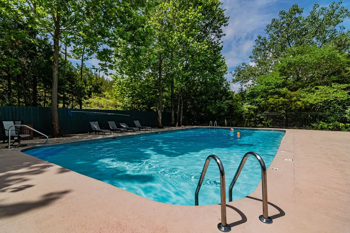 This image showcases a sparkling resort-style swimming pool surrounded by lounge chairs and lush trees, creating an inviting atmosphere for sunbathing, relaxing, and socializing. The clear blue water and natural setting hint at mountain vistas nearby, enhancing the sense of a peaceful, scenic escape.