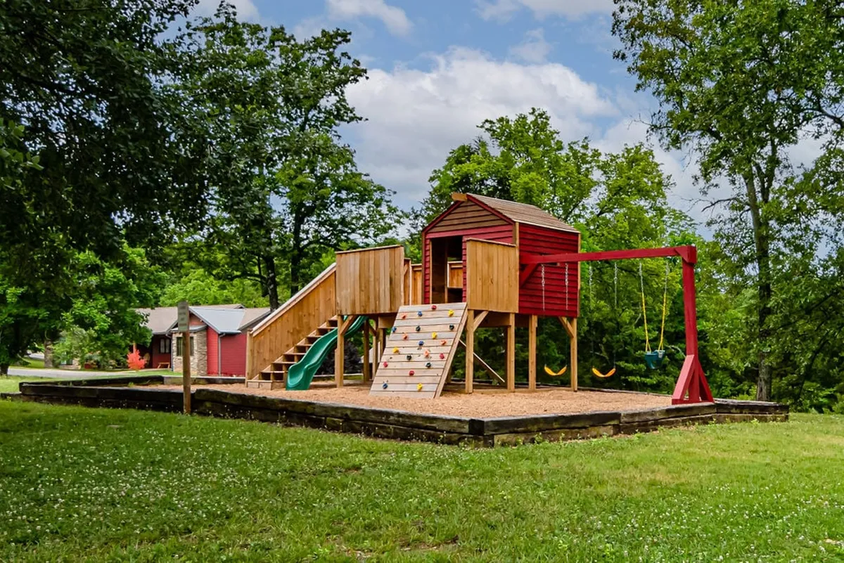 A vibrant playground structure featuring a slide and swing set, surrounded by green trees, designed for children’s outdoor play and exploration. The area is spacious, accommodating multiple families and inviting group activities.