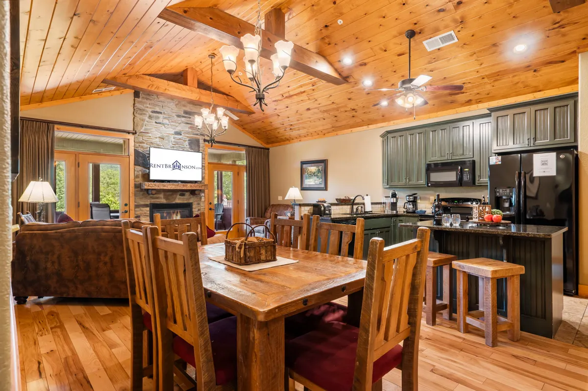 A spacious, inviting open-concept living and dining area featuring a large wooden dining table, a cozy stone fireplace, a chandelier overhead, and expansive windows that flood the room with natural light. The kitchen area is seamlessly integrated into the space, ideal for both meal preparation and entertaining.