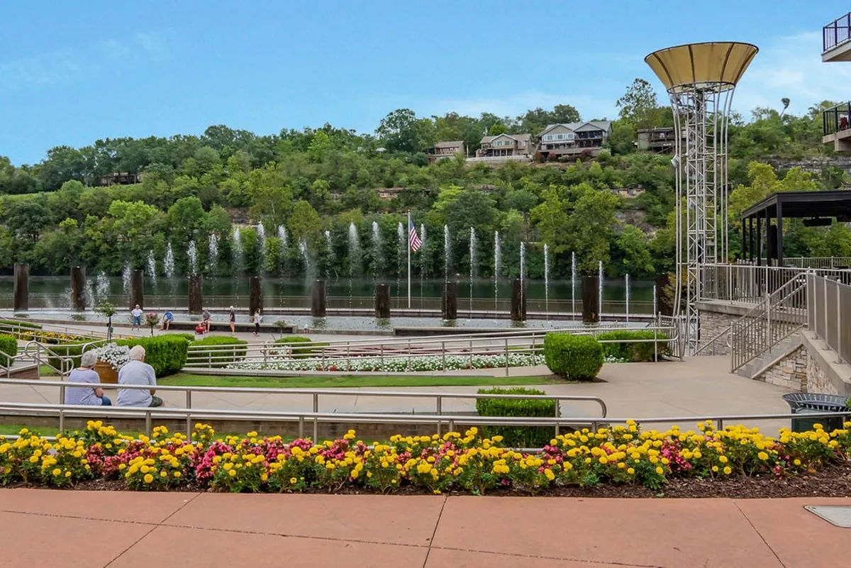 A vibrant exterior scene with flower beds, a water fountain, ample seating, river views, and abundant greenery. Guests can imagine strolling, relaxing, or attending events within this lush setting, which remains fully open for enjoyment.