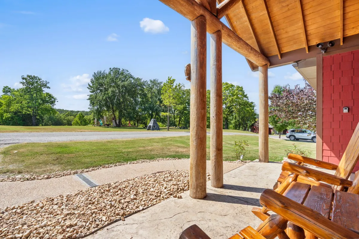 An exterior shot featuring log pillars, a rock path, classic wooden rocking chairs, and lush green trees. The gravel road and natural surroundings emphasize the property’s tranquil setting, providing a broader perspective on the accessible and enjoyable amenities.