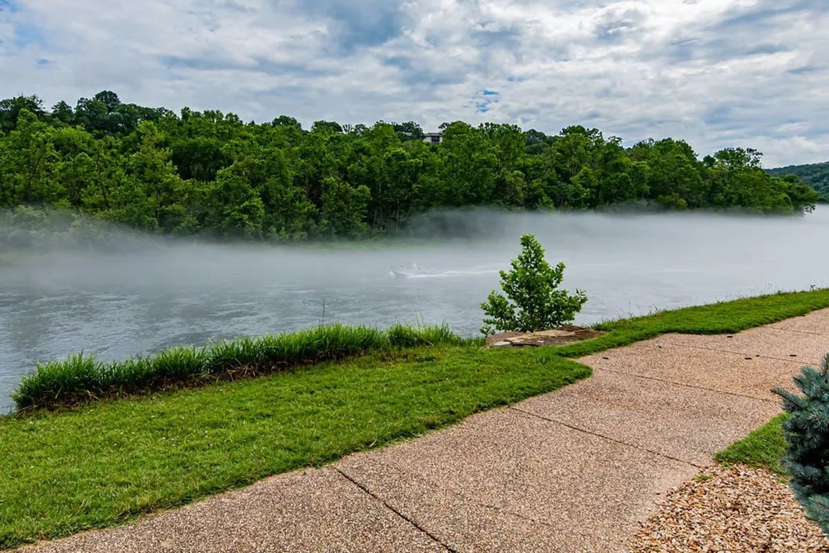 This serene view features a foggy river winding through lush green trees, with a clear pathway along the riverbank. The scene evokes tranquility and offers a glimpse of outdoor adventure opportunities like fishing, walking, and nature observation.