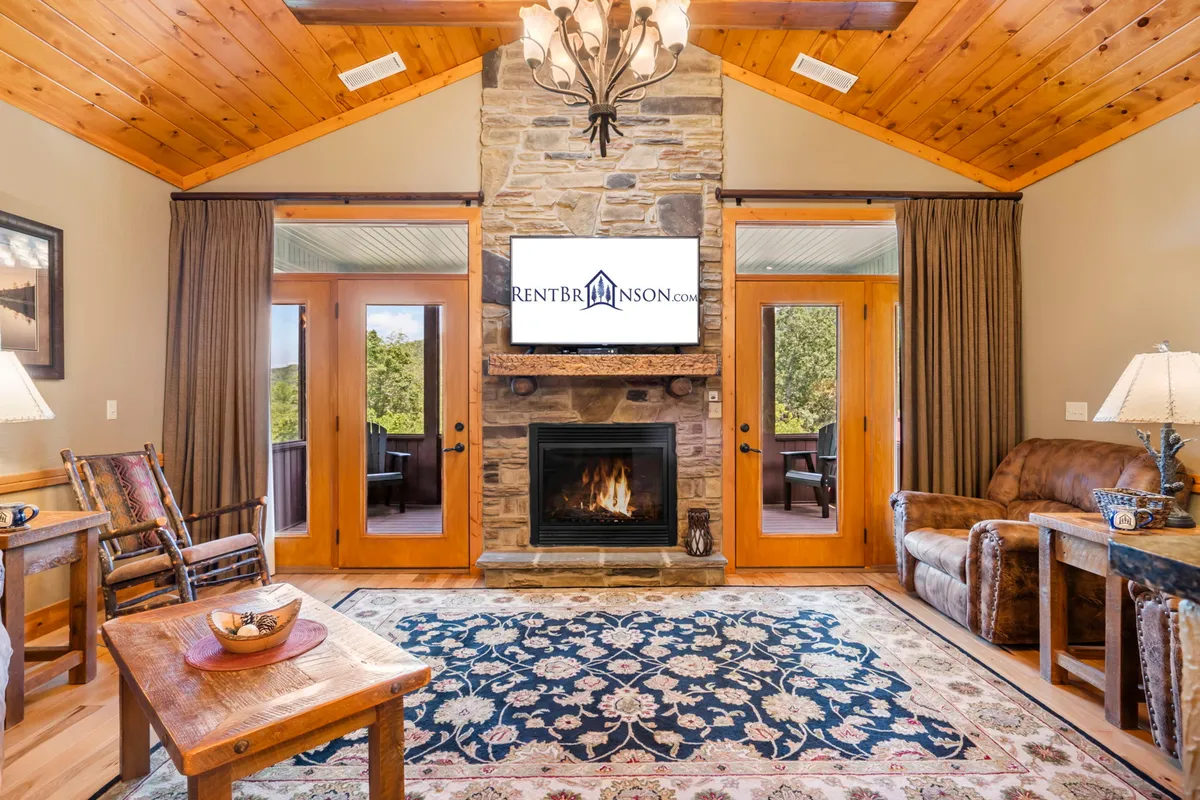 The image showcases a warm, inviting living area with a striking stone fireplace as the centerpiece, high wooden ceilings, large windows revealing glimpses of the Ozarks outdoors, plush and comfortable seating, and a decorative rug that adds a cozy touch. The setup is perfect for gatherings, relaxation, and enjoying the beautiful natural light and views.