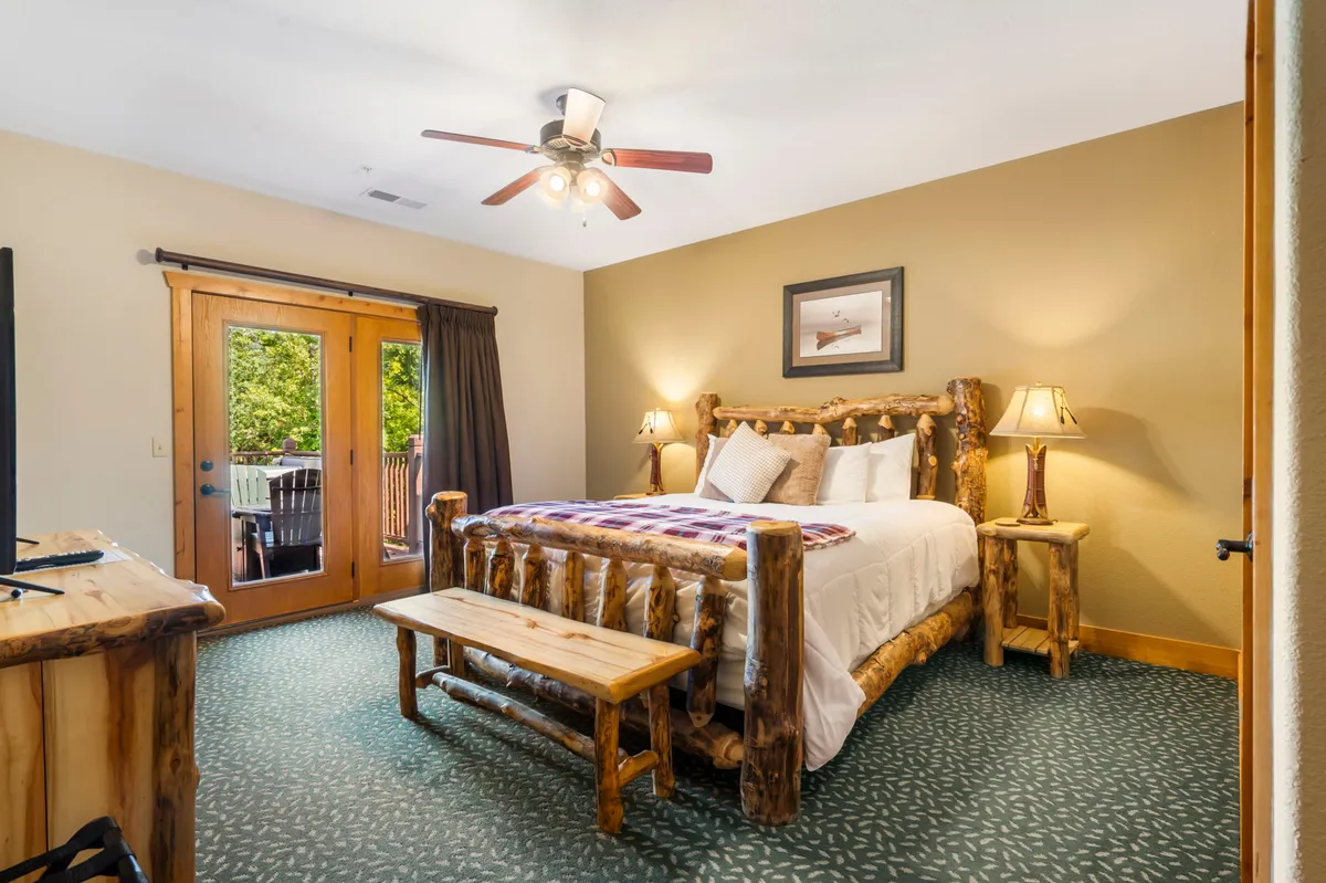 A master bedroom with a rustic log bed, wooden furniture, decorative lamps, and patio doors opening out to a deck. Sunlight streams in, and the warm decor provides an inviting sanctuary for relaxation and nature appreciation.