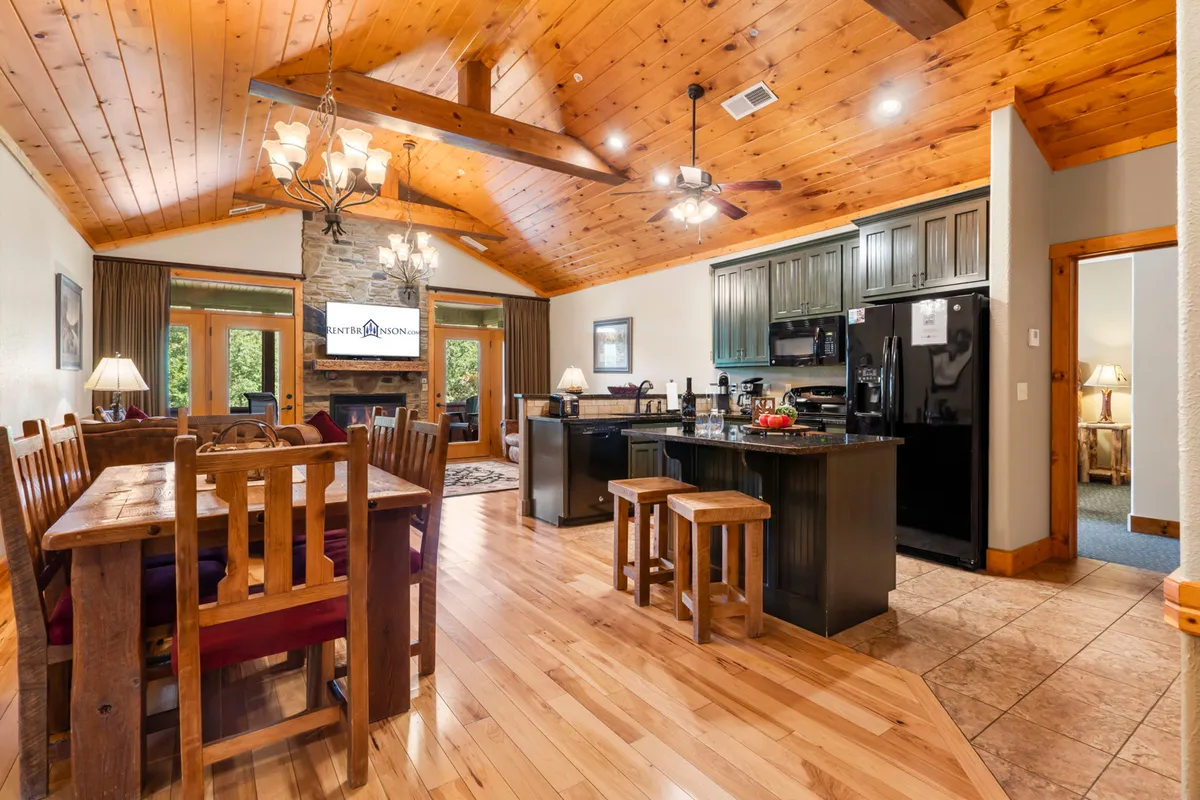 An open living area with a central stone fireplace, a large wooden dining table, a kitchen island, and generous windows overlooking the outdoors. The room is styled with cozy Adirondack-inspired furnishings and hardwood floors, fostering a welcoming atmosphere for family gatherings and relaxation.