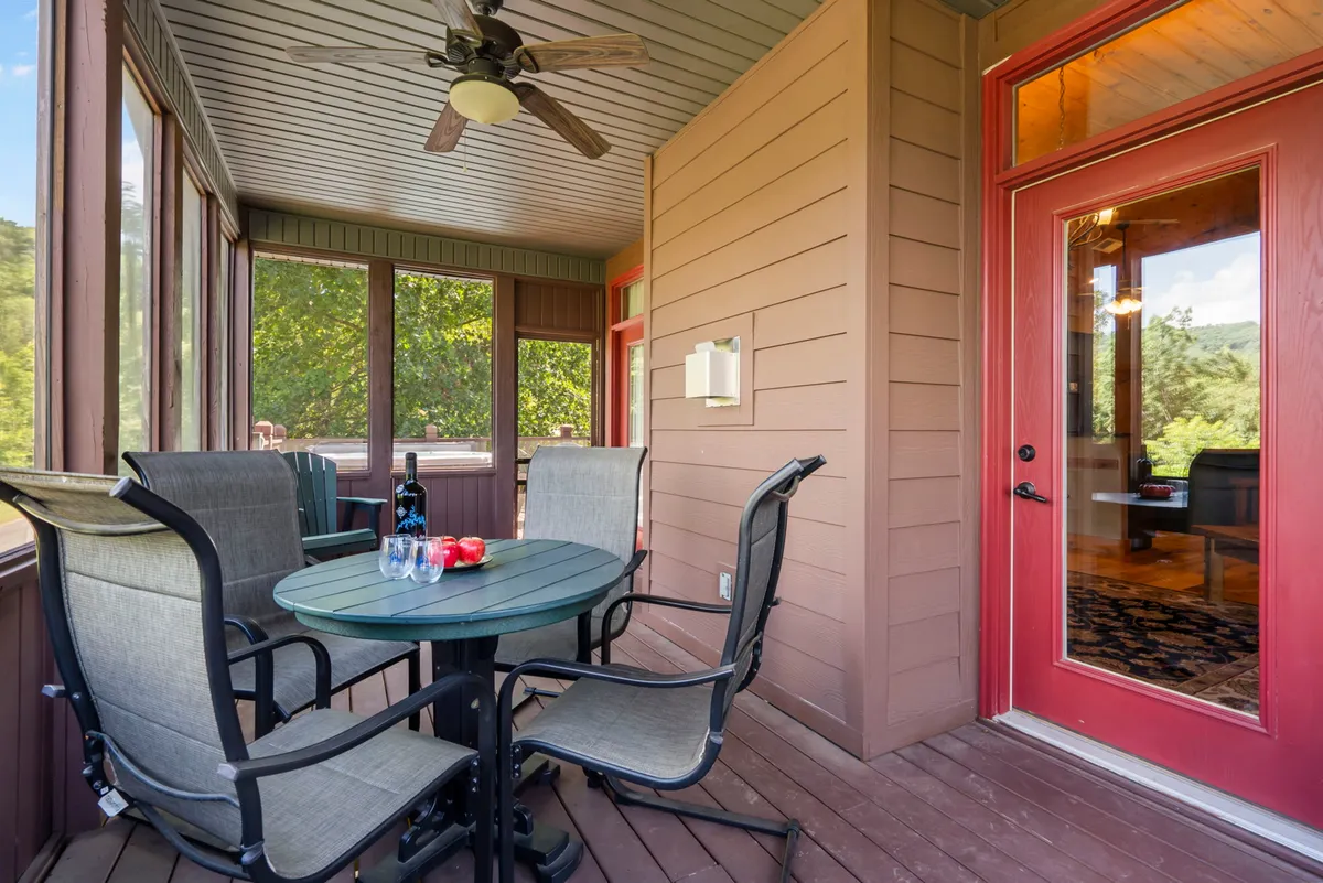 An outdoor seating area on a charming porch featuring a dining table, a glass of wine, fresh tomatoes, and overhead ceiling fan. Surrounded by greenery, this space hints at private alfresco meals and drinks while enjoying nature's tranquility.