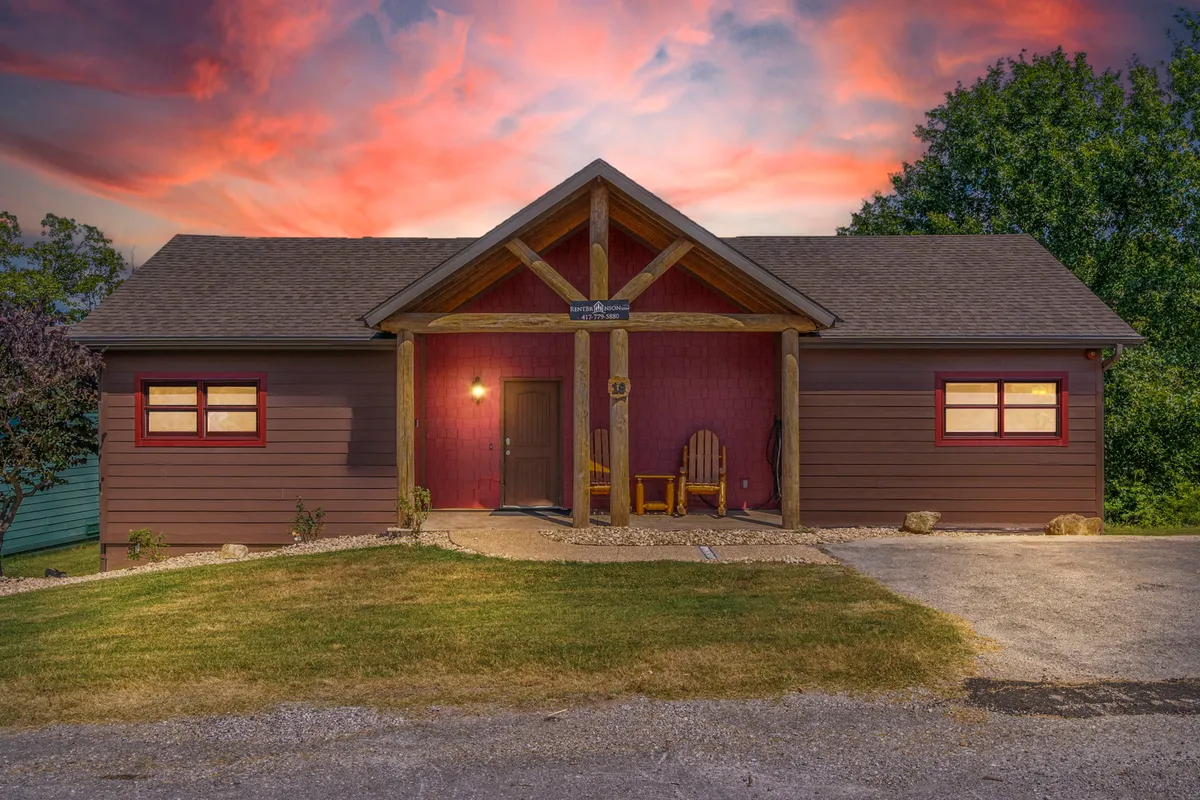 A welcoming front porch with wooden rocking chairs set against a decorative front door, bathed in the warm glow of a colorful sunset. The inviting seating area suggests a place to relax and enjoy each other’s company while taking in the natural beauty around.